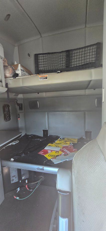 Kenworth T680 truck cab interior showing upper sleeper shelf with netting, lower storage area with scattered paperwork and oil/def bags, dashboard area partially visible.