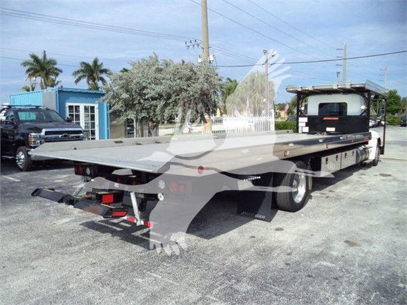 Kenworth T370 tow truck with a long flatbed trailer in a paved lot. Rear view shows flatbed and ramp; cab at front with side mirrors.