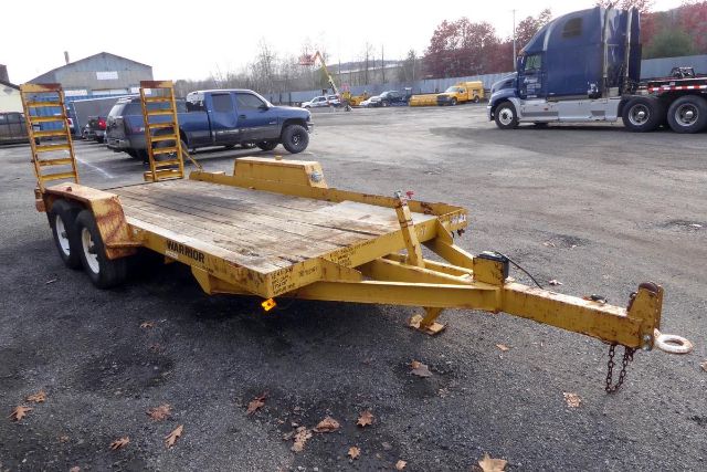 Trailer with dual rear axles and a flatbed, yellow in color, hitch and chains at the front, viewed in an outdoor lot with other vehicles in background.