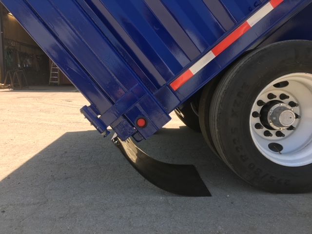 Blue dump trailer with tilting bed raised; rear wheel and mud flap visible; small red marker light; black mudguard; square dump trailer body with reflective orange/white stripe.