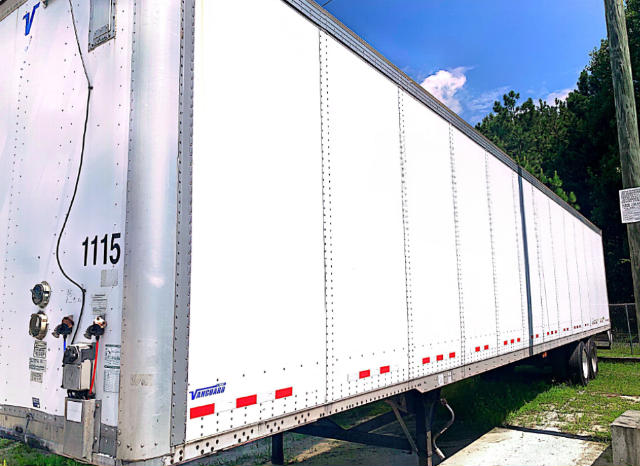 Trailer side view showing white Vanguard trailer with stock number 1115, utility category, VIN 5V8VC5322JM803441, and tandem axles in a grassy lot.