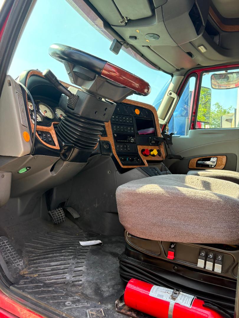 Truck interior showing steering column with black accordion cover, wooden dash trim, multiple gauges, gear selector, center console with switches, gray fabric seat, and red fire extinguisher on the floor.
