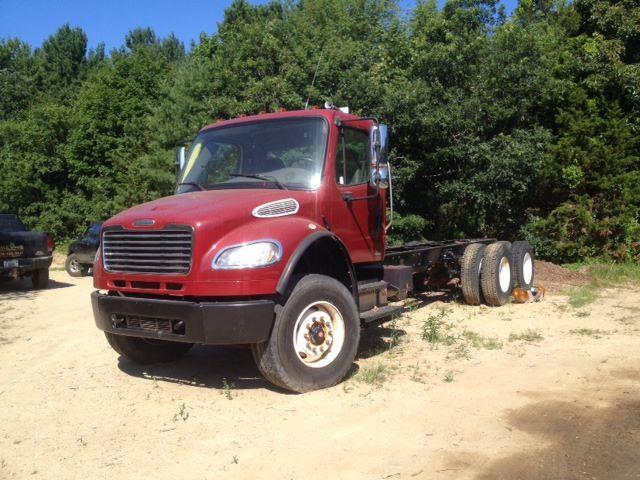 2008 Freightliner M2106 cab chassis truck with red cab, large grille, two front wheels, and dual rear axles on a dirt lot.