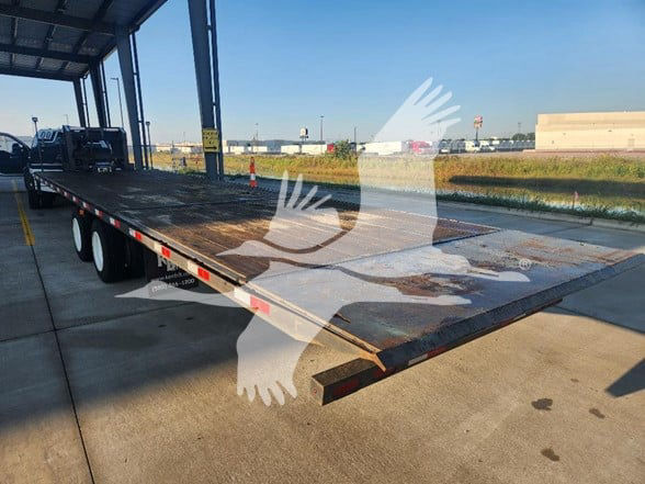 Flatbed trailer showing decking and steel frame with ramp at rear, dual rear axles, outdoor lot in background.