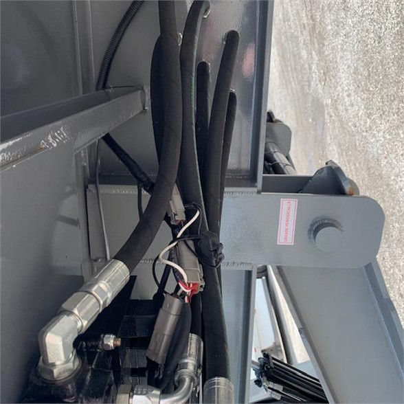 Container delivery trailer hookup area with hydraulic hoses and fittings, metal frame, and a pink warning sticker.
