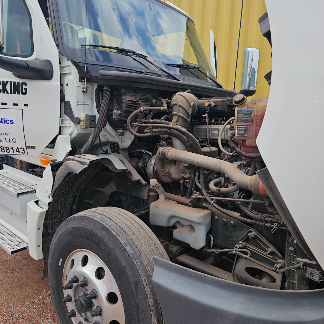 Truck engine bay showing radiator reservoir, hoses, intake, and front wheel; visible label on coolant reservoir and engine components.