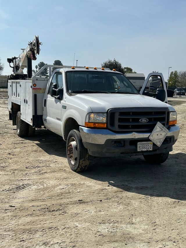 Ford F-550 utility truck with bucket lift on a dirt lot, front grille, license plate visible, side mirrors, and equipment rack in rear.