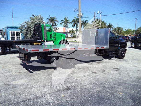 Flatbed truck with green tow vehicle in background, aluminum flatbed and silver cab, roadside lot.
