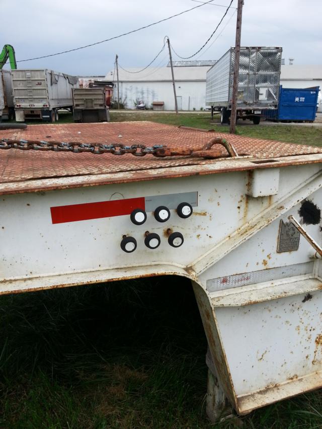 Trailer bed edge with circular valve outlets and a rusted chain latch; white lowboy trailer showing black circular caps and red reflective stripe.