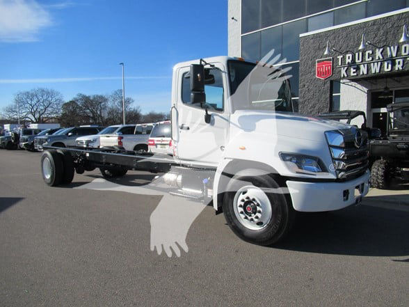 Truck chassis with cab and chassis body, front view showing white daytime running lights and large grille. Text on building reads TRUCKWO KENWOR? but not fully legible.