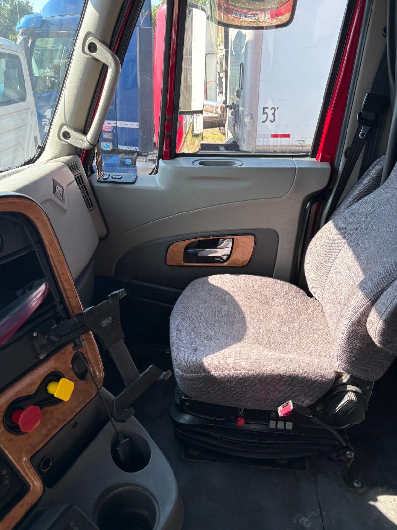 Interior of a 2016 International Prostar truck cabin showing driver seat, grey fabric upholstery, wood trim panel, and dashboard controls with red and yellow knobs.