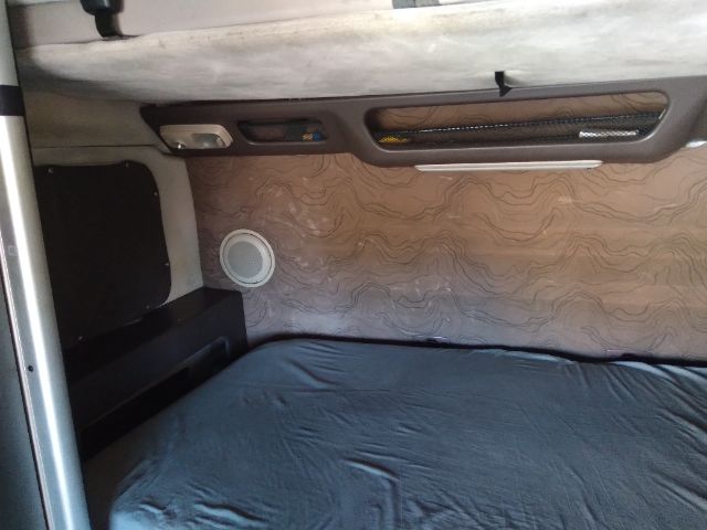 Truck sleeper cabin interior with patterned wall trim and a round air vent, bed with blue sheet, window controls and storage pockets along the wall.