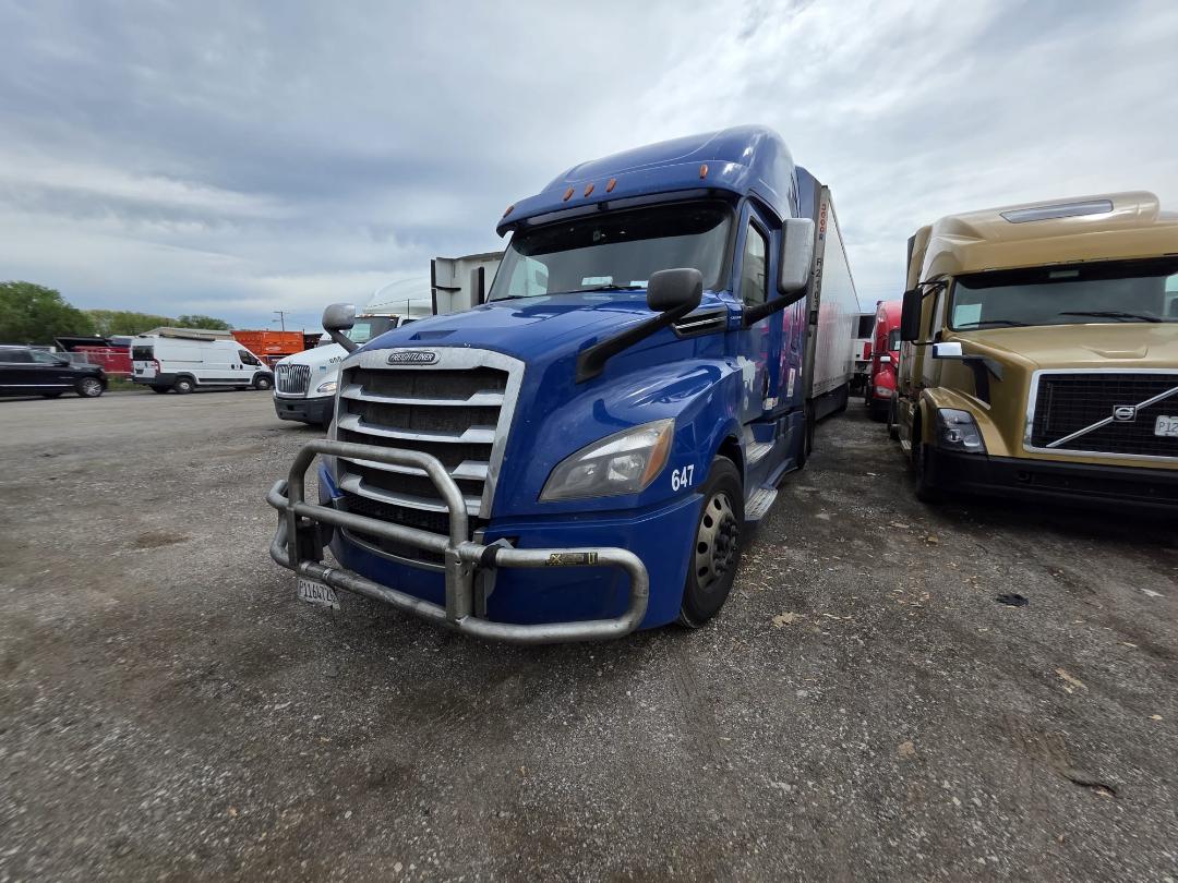 Blue Freightliner Cascadia daytime shot with front bull bar, stock number 647 visible on the fender, parking lot with other trucks.