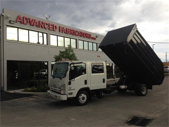 Truck with a raised dump body in front of a building with 'Advanced Fabricators' sign. White cab, black dumping container lifted at an angle.