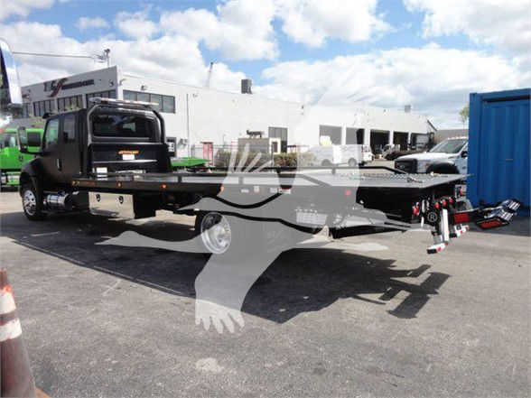 Truck flatbed on a dealership lot with a black cab and extended flatbed, International MV logo visible; trailer equipment and other vehicles in background.