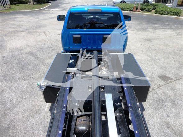 Truck bed/wheel area showing a blue Ram 5500 cab with a heavy-duty towing or flatbed apparatus. Visible components include a metallic platform and hydraulic or mechanical framework, with a dark fuel tank-like cylinder and rails in the foreg