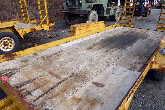 Trailer bed with weathered wooden planks showing wear, undercarriage and yellow frame, with industrial equipment in the background.