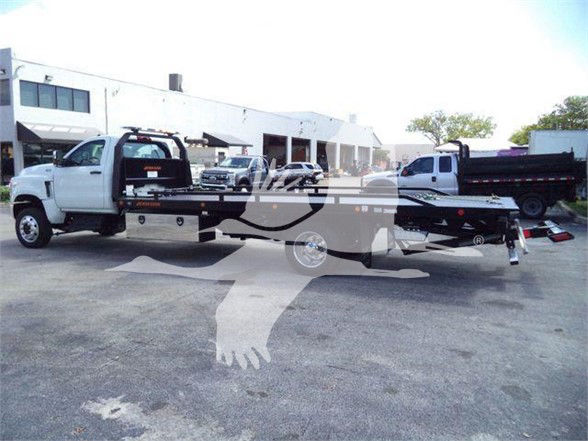 Tow truck flatbed with white cab and long black trailer, dealership lot in background, faint watermark across image.