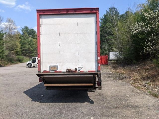 Rear view of a red box truck with a white cargo door and a lowered liftgate, pavement and trees in background.