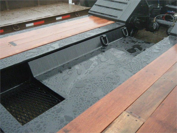 Flatbed trailer deck with wooden boards and a black steel surface. Visible are two metal tie-down points and a mesh storage compartment. Water droplets on the metal surface.