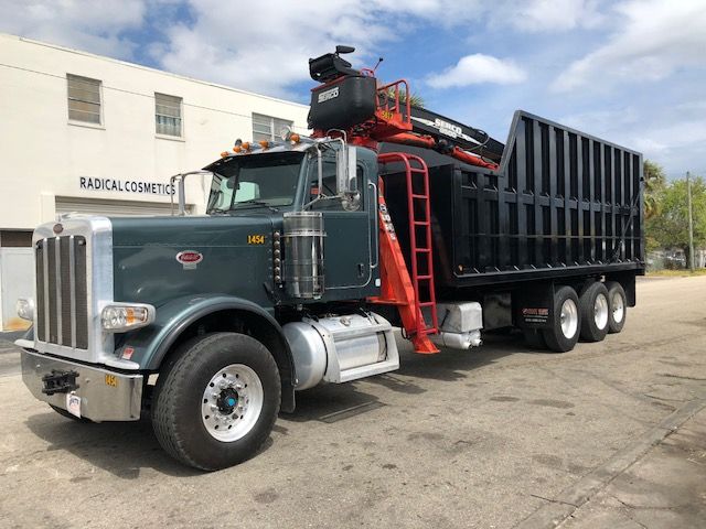 Peterbilt 388 dump/truck with grapple crane on a heavy hauler, side view showing truck cab, black dump bed, red ladder, and crane equipment.