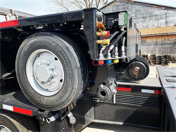 Truck trailer rear with hydraulic hoses, hydraulic manifolds, and spare tire mounted on the side; visible components include hoses, metallic fittings, and a coupling arm.