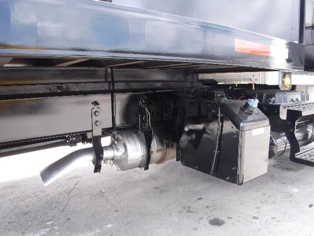 Underbelly of a flatbed truck showing exhaust pipe, drive shaft, and a black rectangular fluid reservoir. Side of truck bed is visible with a blue and white reflective strip.