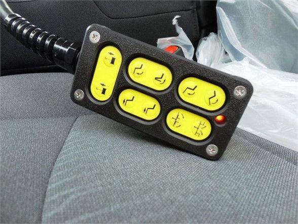 Six-button yellow-illuminated switch panel with icons on each button, mounted on a vehicle seat; visible wiring loom to left; plastic wrap and seat fabric in background.
