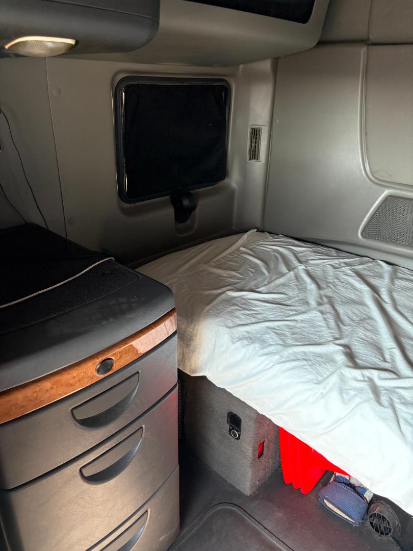 Interior sleeper cab of a 2016 International Prostar showing a bed with white sheet, a wood-trim center console with drawers, and side panel with small window and controls.