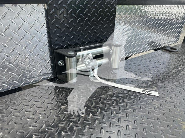 Trailer deck showing tie-down latch and diamond plate surface with RC-branded strap. Flatbed trailer detail near step edge.