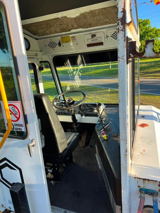 Truck cab interior with steering wheel, dashboard gauges, and black driver’s seat; white exterior with rust on door frame; window view of road.