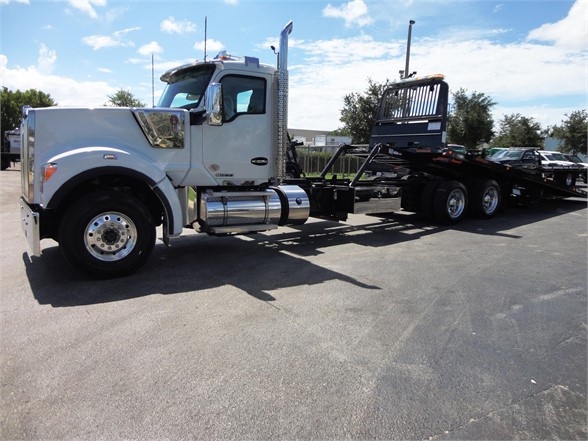 Kenworth W900 truck with a long wheelbase and a flatbed trailer, gray cab, chrome fuel tanks, and a heavy-duty winch in a dealership lot.