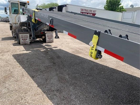 Container delivery trailer with a long gray drop deck extending from a truck cab, yellow straps, red and white reflective markings, and a tan tandem axle configuration.