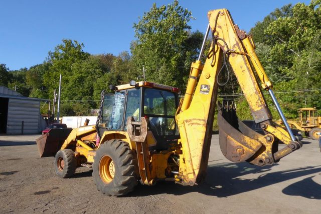 Backhoe loader in operation with front loader and hydraulic arm extended, yellow John Deere machine.