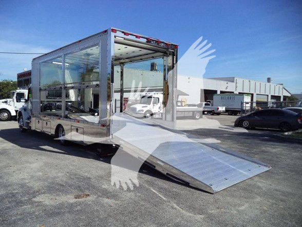 Truck body in a dealership lot with a rear ramp extended; large windows along the sides; chrome exterior; typical Kenworth-style cab visible in background.