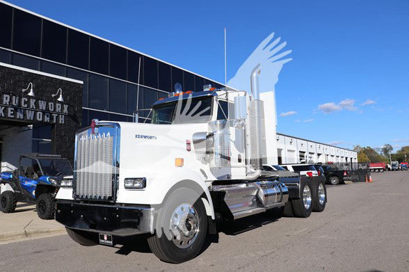 Kenworth W900L conventional daycab truck in front of dealership building. Prominent chrome grille, large vertical exhaust stacks, dual rear axles, white cab with Kenworth badge, truck dealership signage in background.