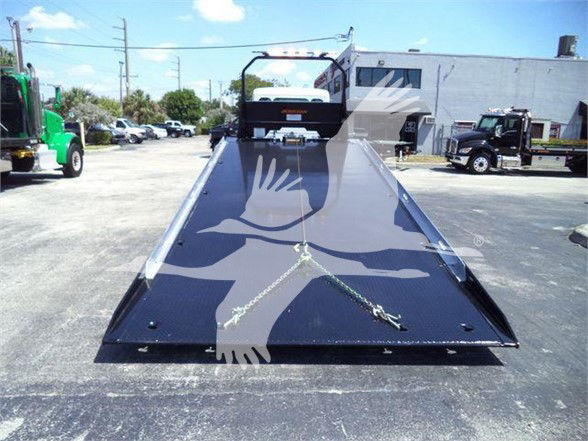 Tow truck with a flatbed carrier; front view showing the extended black ramp bed and wheel chocks, Kenworth cab in background.