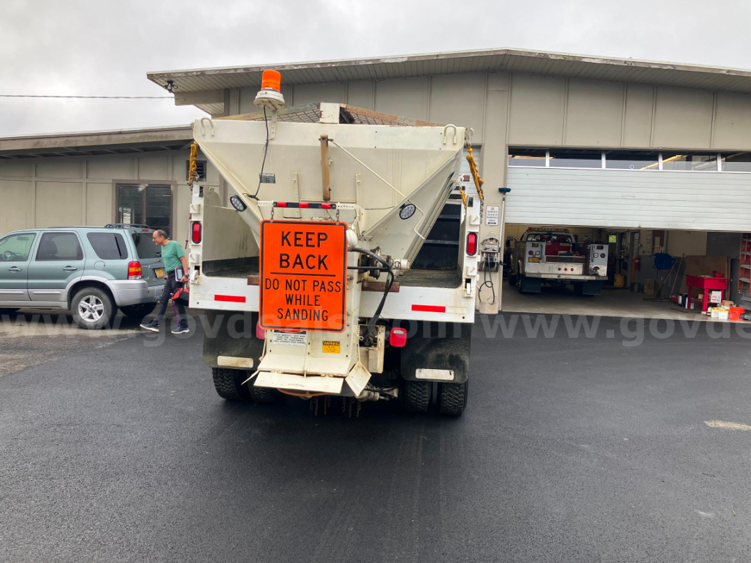 Rear view of a white Ford F-7000 dump truck with an orange 'KEEP BACK' sign on the back and an amber beacon light on top. The truck is in a service area.