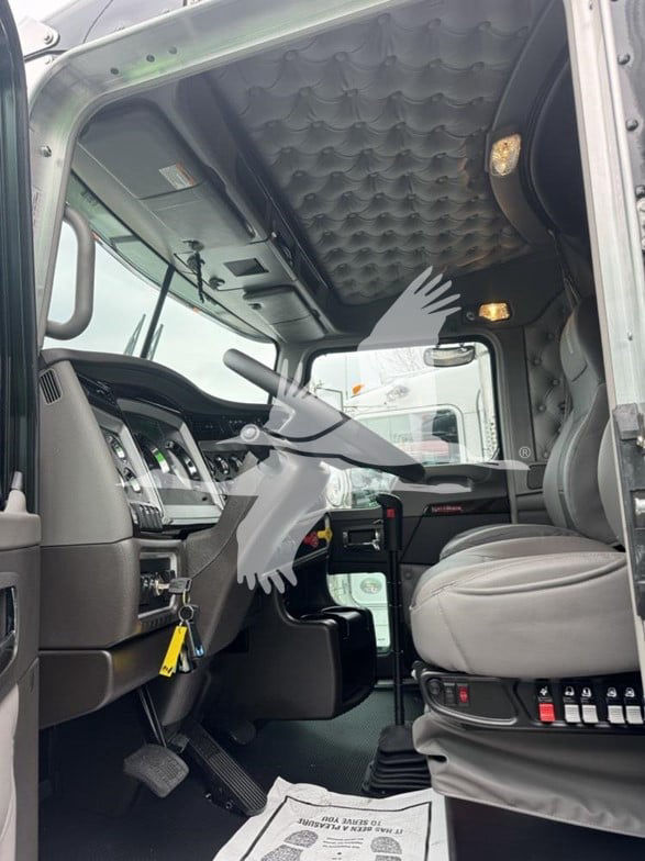 Kenworth W900L truck interior showing driver seat, dashboard, and quilted overhead sleeper area. Steering wheel, pedals, control switches, and sun visor visible.