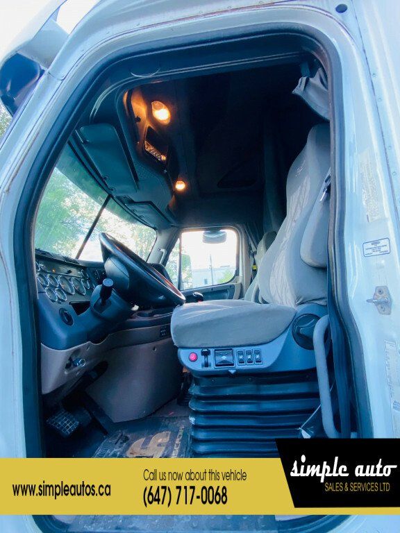 Cab interior of a 2012 Freightliner Cascadia truck showing driver seat, steering wheel, dashboard, and entry with overhead lights.