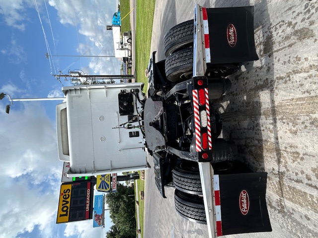 Rear view of a white 2020 Peterbilt 579 tractor chassis with dual rear axles, mud flaps labeled Peterbilt, and a striped rear underrun protection bar.