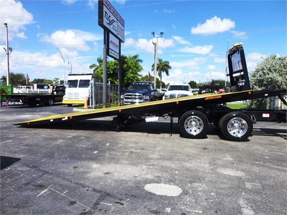 2022 Kenworth W900 tow truck with a long flatbed trailer and dual rear wheels. The angled bed is extended outward. A signboard and other vehicles are in the background.