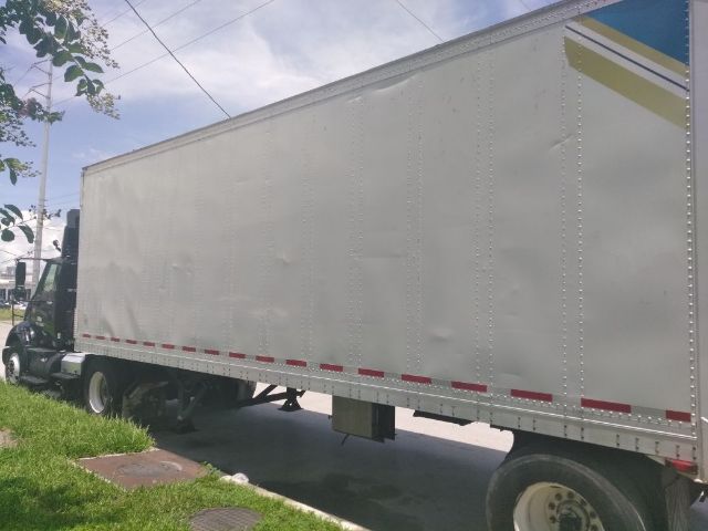 Box trailer side view with cab of semi-truck at left, white aluminum side panels, red reflective tape along lower edge, blue and yellow graphics at rear corner.