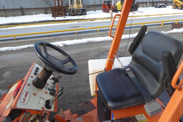 Trucking equipment cab with steering wheel and control panel, orange frame, black seat, snow on ground.