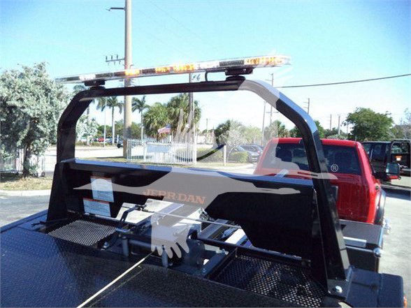 Tow truck bed with metal rack and orange warning light bar; back view shows a Ram logo on the tailgate and a platform with perforated metal; a truck bed with equipment and a red pickup behind.