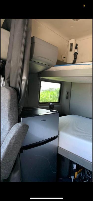 Truck cab interior showing sleeper area with a small fridge, bunk bed, and window.