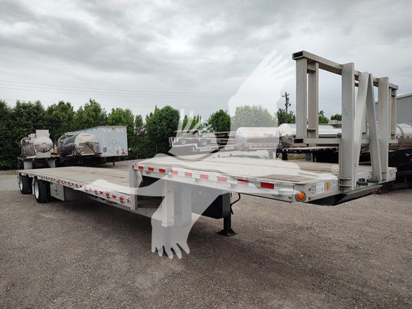 Flatbed trailer with drop deck and gooseneck-style front, no visible brand markings on deck surface. Parking lot with other equipment in background.