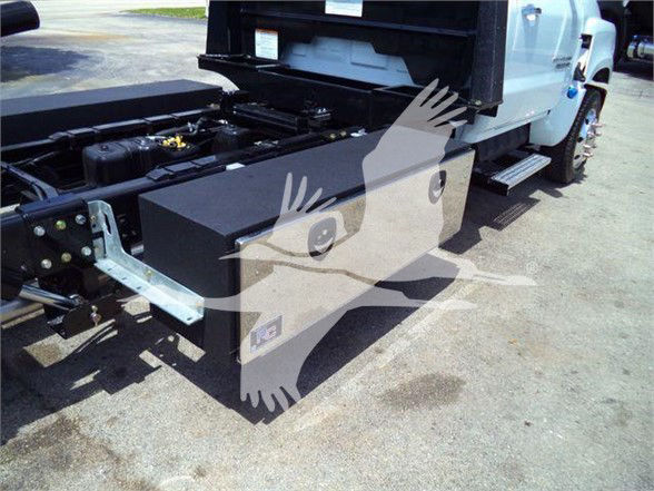 Truck chassis with large black toolbox mounted near front wheel, white cab, hydraulic lines, and metal frame visible.