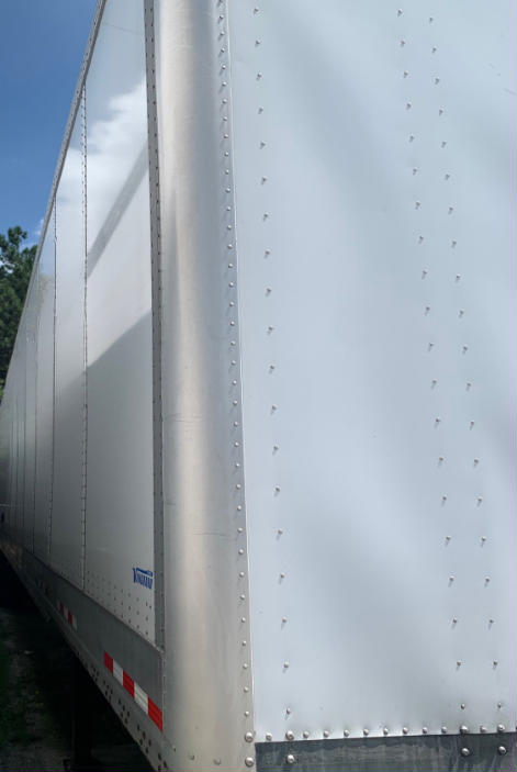 Trailership side of a 2018 Vanguard VXP trailer, riveted aluminum panels, blue Vanguard logo near lower left, reflective red/white stripes at bottom edge.