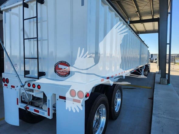 Trailer rear and side view showing a white hopper trailer with a ladder on the left, rear lights, and a decal of hands overlapping. Visible round black and red badge on the back near the left taillight area.